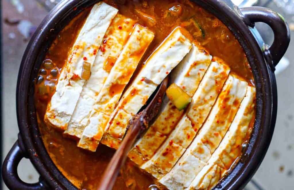 Close-up of a traditional Korean stew in a black pot, featuring thick slices of tofu immersed in a rich, spicy red broth. The soup contains visible ingredients such as sliced leeks and chili flakes. Steam rises, indicating its hot and freshly made.